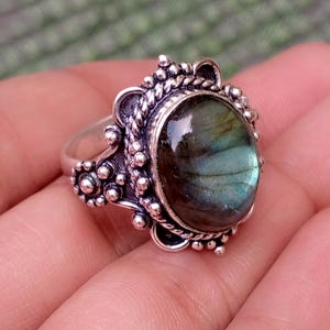 May include: A silver ring featuring an oval-shaped labradorite gemstone with iridescent blue and green flashes. The setting is crafted with intricate metalwork, including a twisted rope border and small decorative silver spheres around the stone.