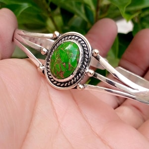 May include: A silver cuff bracelet featuring a central oval green stone with brown veining. The stone is set in a decorative twisted rope border with small silver beads at the cardinal points, attached to a split-shank band design.