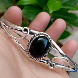 Peut inclure: Un bracelet manchette en argent orné d'une pierre noire ovale centrale sertie dans une bordure en corde torsadée. La bande métallique présente des volutes décoratives de chaque côté de la pierre, créant un design ouvert qui s'affine.