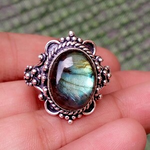 May include: An oval-shaped labradorite gemstone ring set in a decorative silver metal band. The stone displays iridescent blue and green flashes within its dark, polished surface. The silver setting features a twisted rope border and small beads.