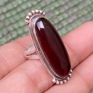 May include: A silver ring featuring a long, oval-shaped, deep red gemstone set vertically. The metal band has a polished finish with decorative beaded detailing at the top and bottom edges of the stone setting, creating a distinct frame.