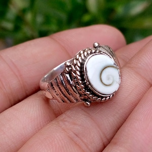 May include: A silver ring featuring a central oval-shaped white shell inlay with a natural brown spiral pattern. The metal band is decorated with a textured rope-like border and horizontal grooved lines along the sides of the setting.