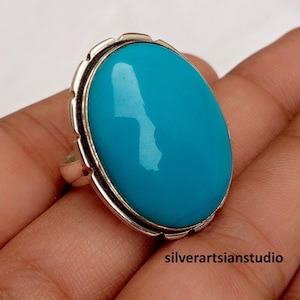 May include: A silver ring featuring a large, smooth, oval-shaped turquoise stone set in a polished metal bezel. The setting has a scalloped edge detail around the perimeter of the stone. The text silverartsianstudio is visible in the corner.
