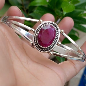 Puede incluir: Una pulsera de plata con una piedra preciosa central de color rosa intenso, forma ovalada y tallada en facetas. La piedra está engastada en un borde de estilo cuerda trenzada y montada sobre una banda que se divide en tres hilos de plata.