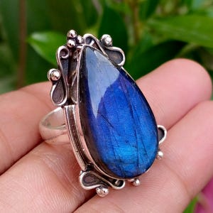 Puede incluir: Un anillo de plata con una gran piedra preciosa de labradorita en forma de pera con un vibrante destello azul iridiscente. El engaste de metal presenta volutas decorativas y pequeñas esferas de plata que rodean la piedra central.