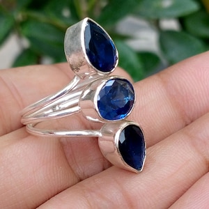 May include: A silver ring featuring three deep blue faceted gemstones. The design includes one oval-shaped stone positioned between two pear-shaped stones, all set in polished silver metal bands that wrap around the finger in a layered arrangement.