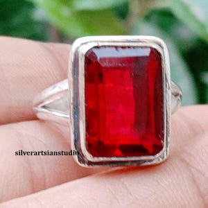 Puede incluir: Un anillo de plata con una gran piedra preciosa roja oscura rectangular y facetada, engastada en un bisel de metal liso. La banda tiene un diseño dividido en los lados. El texto silverartsianstudio está impreso en letras negras en el lateral.