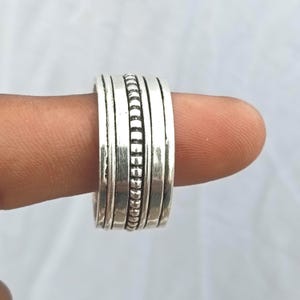 May include: A wide silver-toned ring featuring a multi-band design. The center band has a textured, beaded pattern, flanked by two smooth, thinner bands on each side, creating a layered appearance with a polished metallic finish.