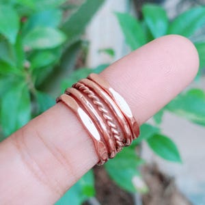 May include: A set of four copper-coloured rings stacked together on a finger. The set includes two smooth, flat bands, one band with a hammered texture, and one central band featuring a twisted rope design, all with a polished metallic finish.
