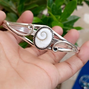 Può includere: Un bracciale rigido in argento con al centro una conchiglia occhio di shiva bianca di forma ovale con un motivo a spirale naturale. La fascia in metallo ha una finitura lucida con dettagli in filo intrecciato su entrambi i lati.