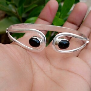 Peut inclure: Un bracelet manchette en argent au design ouvert, orné de deux pierres noires ovales, lisses et polies, fixées à chaque extrémité. La bande métallique présente une surface arrondie et réfléchissante qui s'incurve autour des pierres.