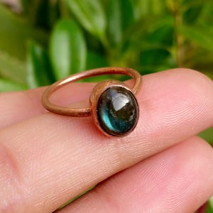 May include: A ring featuring a smooth, polished copper band and a single oval-shaped cabochon gemstone. The stone displays a dark, iridescent blue and green colour with a reflective surface, set securely within a simple copper bezel setting.