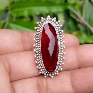 May include: A silver ring featuring a large, elongated, faceted deep red gemstone set in the center. The metal frame surrounding the stone is decorated with a textured, beaded border that creates a sunburst effect around the oval-shaped center.