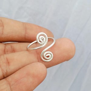 May include: A silver-toned bypass ring featuring two open-ended spiral motifs at each end of the band. The metal has a smooth, polished finish, and the design consists of a continuous wire that curves into circular coils on both sides of the ring.
