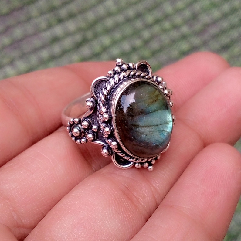 May include: A silver ring featuring an oval-shaped labradorite gemstone with iridescent blue and green flashes. The setting is crafted with intricate metalwork, including a twisted rope border and small decorative silver spheres around the stone.