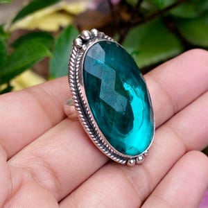 Peut inclure: Une grande bague ovale avec une pierre facettée de couleur sarcelle montée sur une bande en métal argenté. Le sertissage est orné d'une bordure torsadée en forme de corde et de petits accents sphériques sur le bord extérieur du cadre ovale.