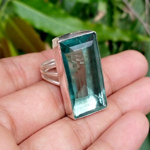 May include: A silver ring featuring a large, rectangular, faceted teal-coloured gemstone set in a smooth metal bezel. The band of the ring is constructed from three thin, parallel silver wires that connect to the base of the gemstone setting.