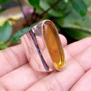 May include: A wide silver ring featuring a large, elongated, faceted amber-coloured gemstone set vertically in a smooth metal bezel. The band has a polished, reflective surface that curves around the finger, showcasing a clean, metallic finish.