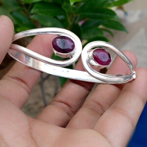 May include: A silver cuff bracelet featuring an open design with two teardrop-shaped terminals. Each terminal is set with a single, faceted, oval-shaped deep red ruby gemstone. The metal band has a smooth, polished finish throughout the structure.