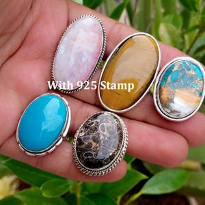 May include: Five oval rings featuring diverse gemstones including a pale pink stone, a golden-brown tiger's eye, a turquoise and copper composite, a solid bright blue stone, and a dark fossilised stone, all set in silver with a 925 stamp.