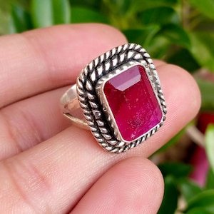 May include: A silver ring featuring a rectangular, faceted, deep pink gemstone set within a twisted rope-style metal border. The band is made of polished silver and the setting has a distinct, textured frame surrounding the central stone.