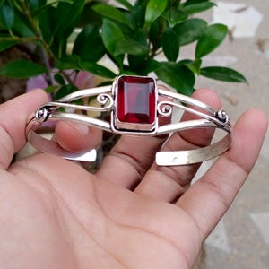 May include: A silver cuff bracelet featuring a central rectangular deep red gemstone. The metal band is constructed with two parallel wires that curve into decorative scrolls on either side of the stone setting, creating an open design.