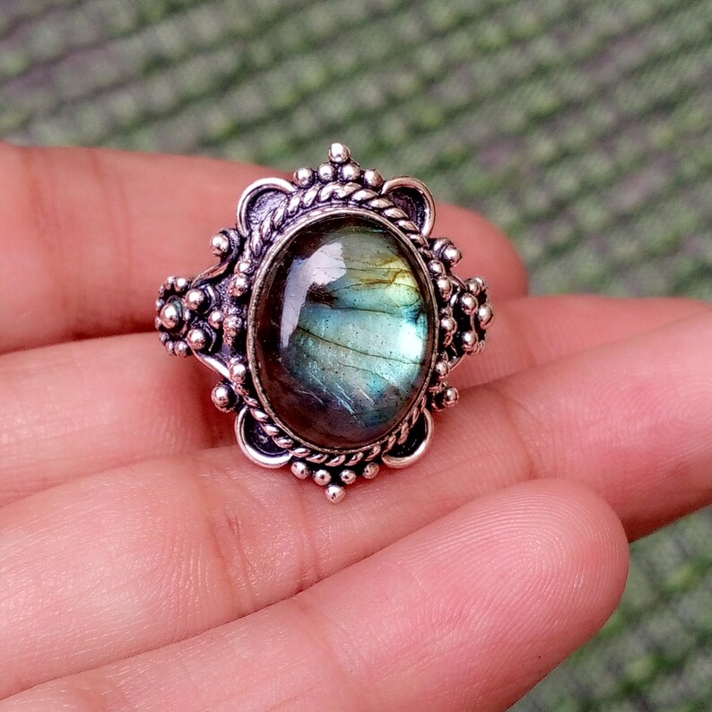May include: An oval-shaped labradorite gemstone ring set in a decorative silver metal band. The stone displays iridescent blue and green flashes within its dark, polished surface. The silver setting features a twisted rope border and small beads.