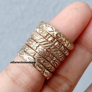 May include: A stack of six gold-toned metal rings, each featuring a distinct embossed pattern including geometric shapes, floral motifs, and textured bands. The text silverartsianstudio is printed in black on the surface of the bottom ring.