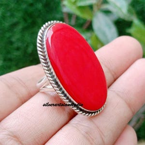 Può includere: Un anello in argento con una grande pietra rossa liscia di forma ovale, incastonata in una cornice con motivo a corda intrecciata. La fascia è in argento lucido e l'incastonatura è rialzata, mettendo in risalto il colore rosso vivido.