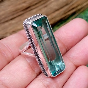 May include: A silver ring featuring a large, rectangular, faceted teal-colored gemstone set in a frame with a twisted rope-like metal border. The band of the ring is smooth and polished, contrasting with the textured detail surrounding the stone.