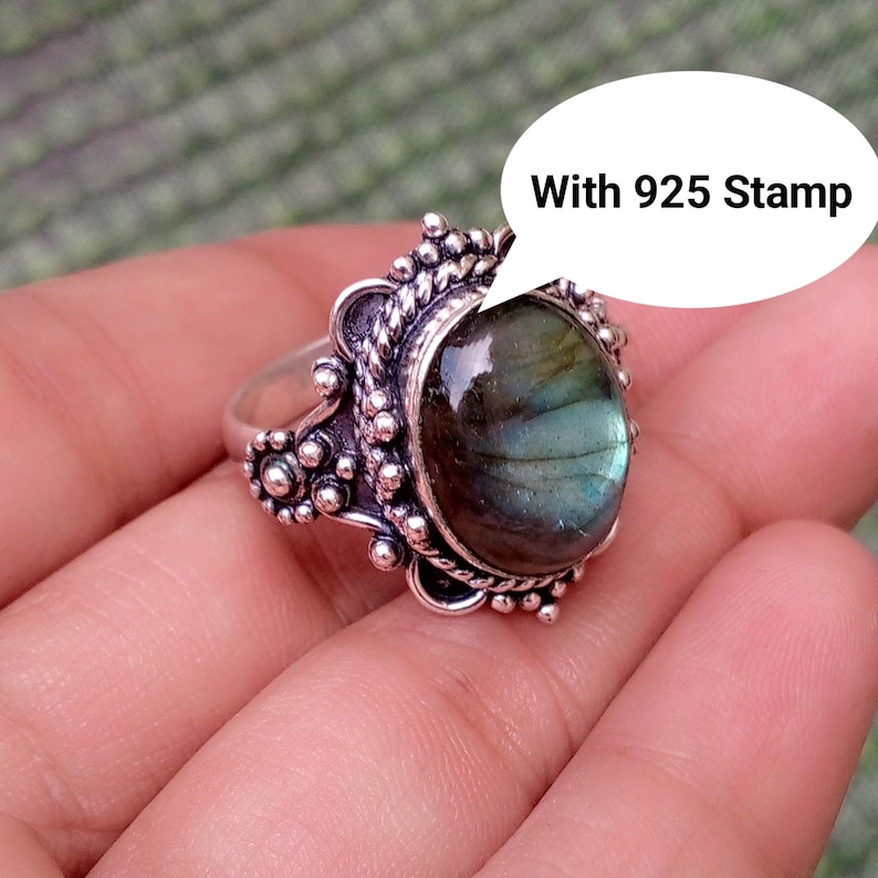 May include: A silver ring featuring an oval labradorite gemstone with iridescent blue and green flashes. The setting is decorated with intricate metal beadwork and twisted wire detailing. A speech bubble graphic includes the text With 925 Stamp.