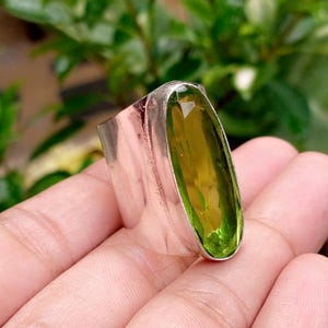 May include: A wide silver band ring featuring a large, vertically oriented, faceted oval peridot gemstone. The stone is a translucent olive green colour, set within a polished metallic frame that wraps around the finger with a smooth finish.