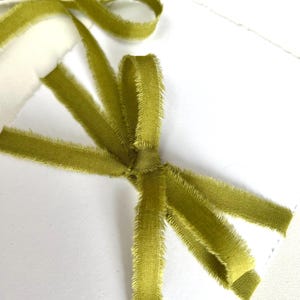 Pode incluir: Uma fita de veludo verde-oliva com bordas desfiadas e cruas, amarrada em um laço simples. A fita apresenta um pelo macio e texturizado, exibida sobre uma superfície branca lisa, destacando a cor verde vibrante e a textura fibrosa delicada.
