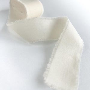Puede incluir: Un carrete de cinta de color blanco roto con una textura tejida suave y delicados bordes deshilachados. La cinta está parcialmente desenrollada, mostrando una longitud continua de tela que se curva a través del marco con un aspecto natural.