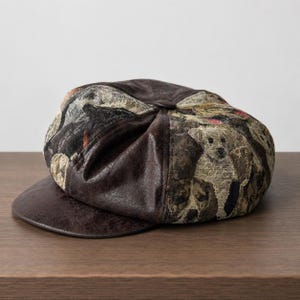 May include: A newsboy cap featuring a dark brown faux leather brim and center panel, contrasted with side panels made of a woven tapestry fabric. The tapestry displays a repeating pattern of various dog breeds in shades of tan, brown, and black.