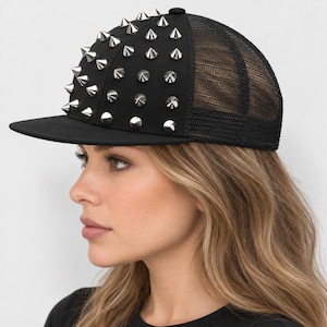 Può includere: Un cappellino da baseball nero con visiera piatta e pannello posteriore in rete. I pannelli anteriori e laterali sono decorati con numerose borchie metalliche color argento disposte a griglia. Il cappellino ha una corona strutturata.