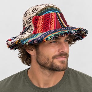 Op de afbeelding: Een bucket hat met een patchworkontwerp, voorzien van een crèmekleurig gedeelte met zwarte paddenstoelillustraties, naast levendige rode, oranje en blauwgroene geweven panelen met geometrische patronen en een gerafelde, veelkleurige rand.