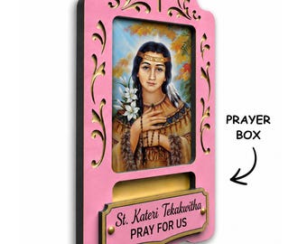 May include: A pink wooden wall plaque featuring a central portrait of St. Kateri Tekakwitha holding white lilies. The frame has decorative cutouts, a cross at the top, and a rectangular plaque below with the text St. Kateri Tekakwitha PRAY FOR US.