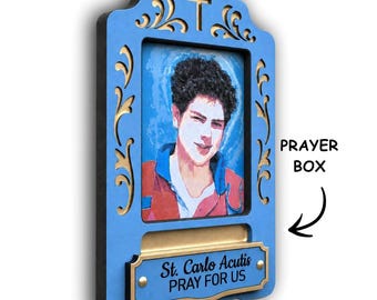 May include: A blue and gold wall-mounted prayer box featuring a portrait of Carlo Acutis. The frame includes a cross cutout at the top and a gold plaque at the bottom with the text St. Carlo Acutis PRAY FOR US. The box has a slot for notes.