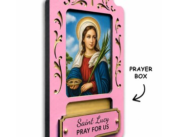 May include: A pink wooden wall plaque featuring a central religious image of Saint Lucy holding a palm branch and a dish with eyes. The frame has a cutout cross at the top and a rectangular slot labelled PRAYER BOX with a plaque reading Saint Lucy PRAY FOR US.