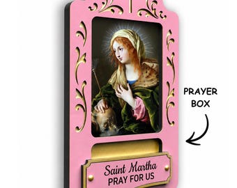 May include: A pink wooden wall-mounted prayer box featuring a central religious portrait of Saint Martha. The frame has decorative cutouts, a cross at the top, and a plaque with the text Saint Martha PRAY FOR US. A slot is provided for notes.