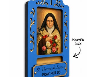 May include: A blue wooden wall plaque featuring a portrait of St. Therese de Lisieux holding a crucifix and roses. The frame has decorative cutouts and a cross at the top. A lower plaque reads St. Therese de Lisieux PRAY FOR US. Text PRAYER BOX.