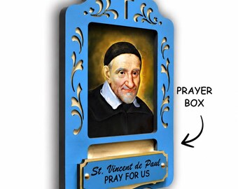 May include: A blue wooden prayer box featuring a portrait of St. Vincent de Paul. The frame has decorative cutouts and a cross at the top. A gold-coloured plaque at the bottom displays the text: St. Vincent de Paul, PRAY FOR US.