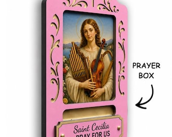 May include: A pink wooden wall-mounted prayer box featuring a cutout cross at the top and an image of Saint Cecilia holding a pipe organ and violin. A rectangular plaque below reads Saint Cecilia PRAY FOR US with a slot for prayer notes.