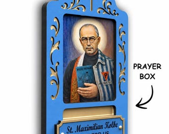May include: A blue wall-mounted prayer box featuring a portrait of St. Maximilian Kolbe in a striped uniform with a halo. The frame has gold filigree, a cross at the top, and a plaque reading St. Maximilian Kolbe PRAY FOR US with a slot.