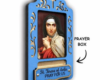 May include: A blue wooden wall-mounted prayer box featuring a central portrait of St. Teresa of Avila with hands clasped in prayer. The frame has gold-painted scrollwork, a cross at the top, and a plaque reading St. Teresa of Avila PRAY FOR US.
