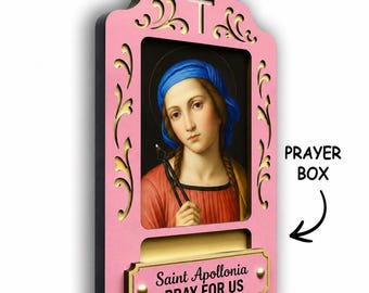 May include: A pink wooden prayer box featuring a central portrait of Saint Apollonia holding dental pliers. The frame has gold-coloured cutouts, a cross at the top, and a plaque at the bottom with the text Saint Apollonia PRAY FOR US.