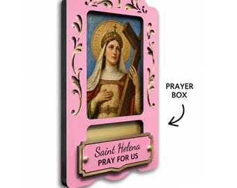May include: A pink wooden wall-mounted prayer box featuring a central religious image of Saint Helena holding a cross. Below the image is a rectangular plaque with the text Saint Helena PRAY FOR US. The frame has decorative cutouts.