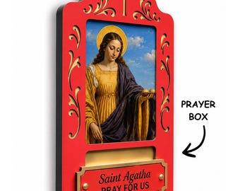 May include: A red and gold wall-mounted prayer box featuring a central image of Saint Agatha holding a plate with two round objects. The frame has decorative cutouts, a cross at the top, and a plaque that reads Saint Agatha PRAY FOR US.