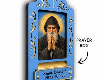 May include: A blue wall-mounted prayer box featuring a central portrait of Saint Charbel with a golden halo, holding a rosary. The frame has decorative cutouts and a plaque at the bottom with the text Saint Charbel PRAY FOR US in black.